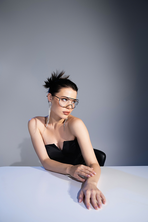 Stylish woman in black corset and sunglasses posing near table on grey background