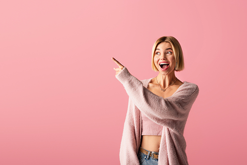 amazed young woman in soft cardigan pointing with finger isolated on pink