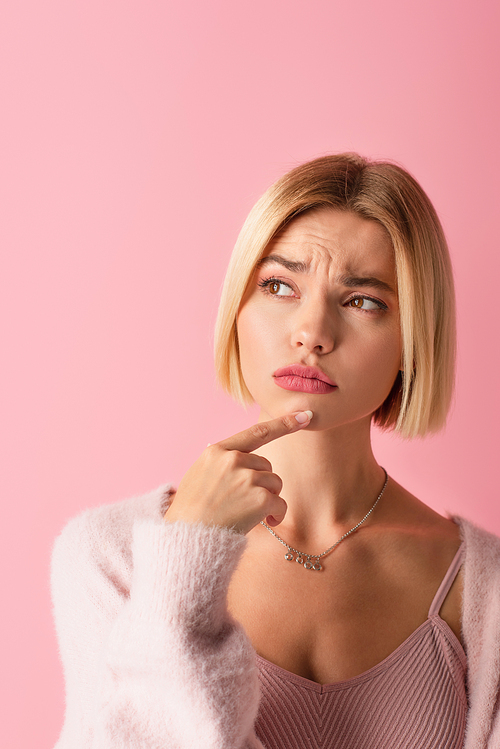 pensive young woman in soft cardigan touching chin with finger isolated on pink