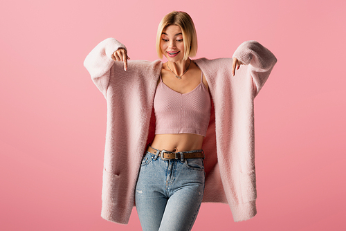 happy young woman in soft cardigan and jeans pointing down with fingers isolated on pink