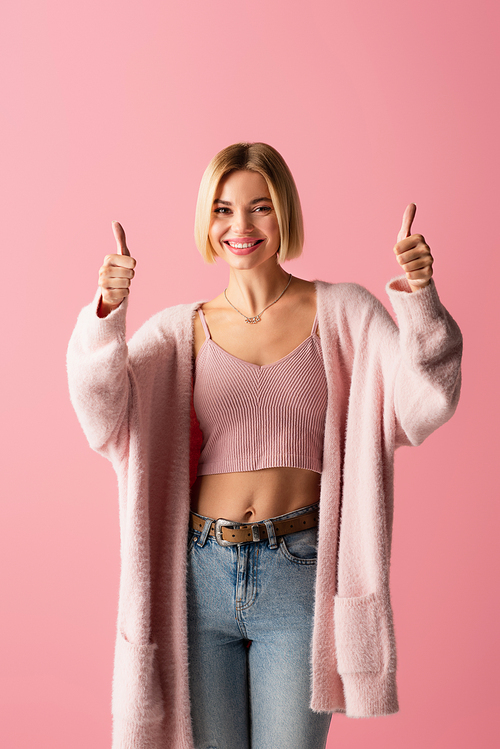 happy young woman in soft cardigan showing thumbs up isolated on pink