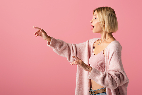 side view of shocked young woman in soft cardigan looking away and gesturing isolated on pink