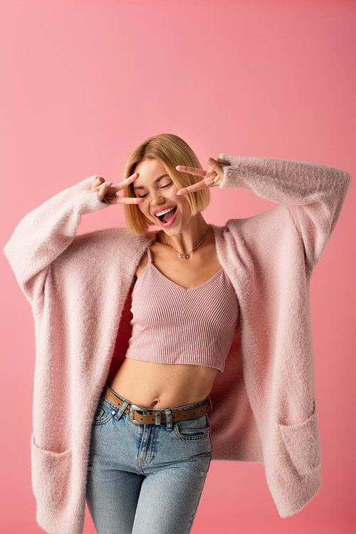 happy young woman in soft cardigan showing peace isolated on pink