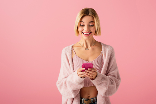 happy young woman in soft cardigan using smartphone isolated on pink