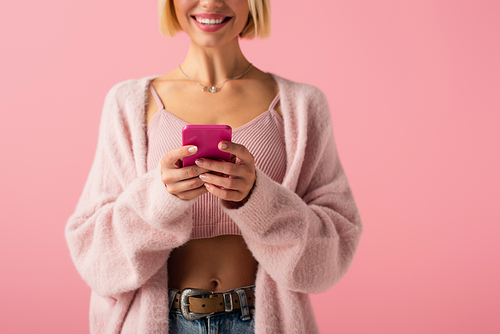 cropped view of happy young woman in soft cardigan using smartphone isolated on pink
