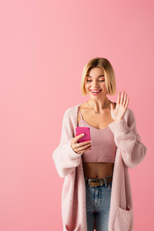 happy young woman in soft cardigan waving hand during video call isolated on pink