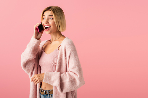 amazed young woman in soft cardigan talking on smartphone isolated on pink