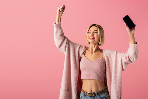 happy young woman in soft cardigan holding smartphone with blank screen isolated on pink