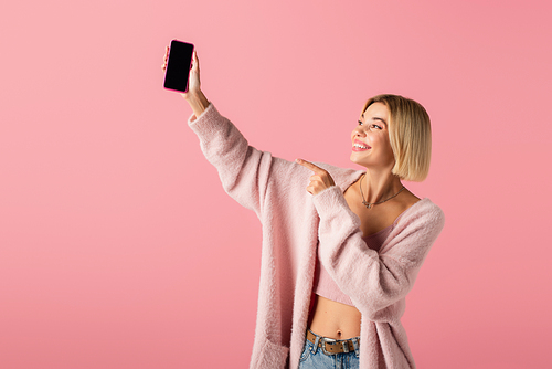 happy young woman in soft cardigan pointing at smartphone with blank screen isolated on pink
