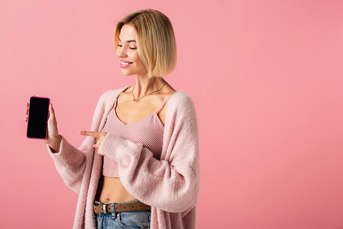cheerful young woman in soft cardigan pointing at smartphone with blank screen isolated on pink