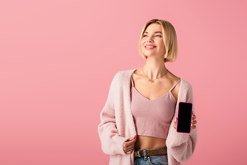positive young woman in soft cardigan holding smartphone with blank screen isolated on pink