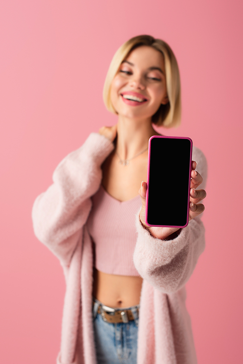 blurred and happy young woman in soft cardigan holding smartphone with blank screen isolated on pink