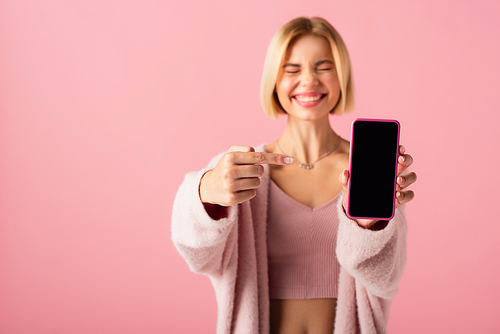 blurred and happy young woman in soft cardigan pointing at smartphone with blank screen isolated on pink