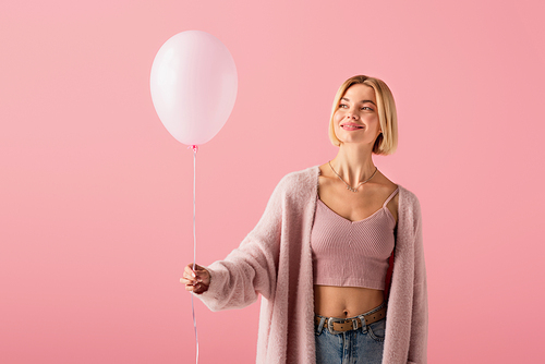 cheerful blonde woman in cardigan holding balloon isolated on pink