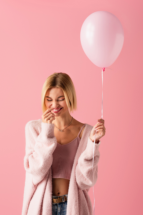 positive young woman in soft cardigan holding balloon isolated on pink