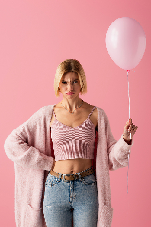dissatisfied young woman in cardigan holding balloon and standing with hand on hip isolated on pink