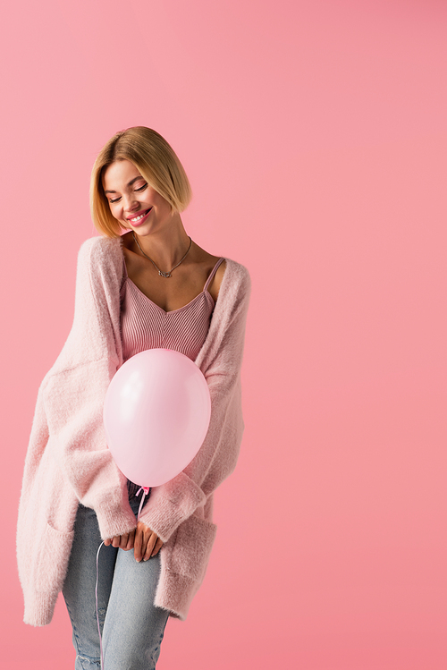 cheerful young woman in cardigan holding balloon isolated on pink