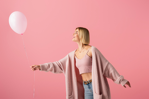 cheerful young woman in cardigan looking at balloon isolated on pink