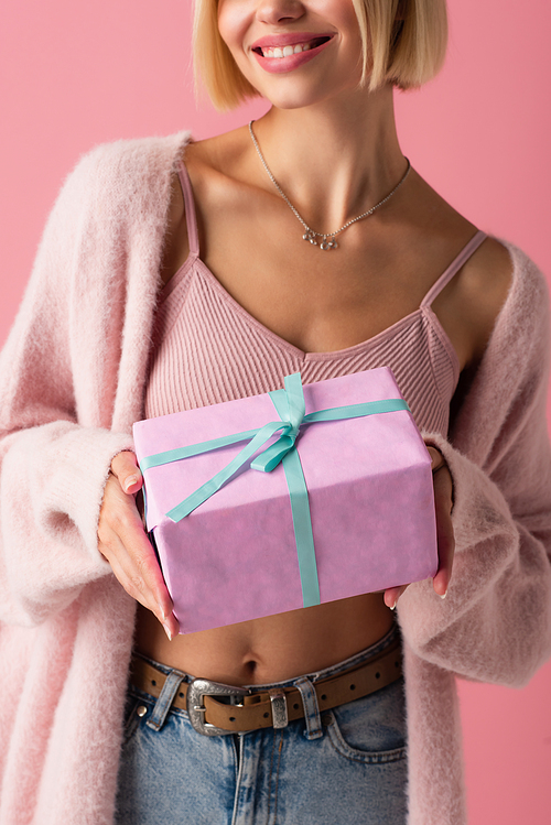 cropped view of cheerful young woman in cardigan holding wrapped gift box isolated on pink