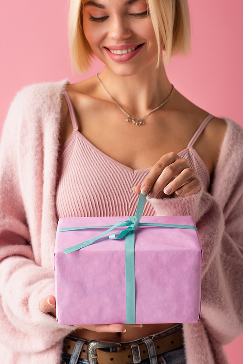 cheerful young woman in cardigan pulling blue ribbon on wrapped gift box isolated on pink