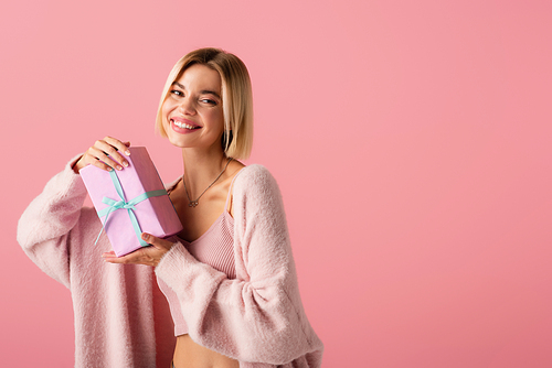 cheerful young woman in cardigan holding wrapped gift box isolated on pink