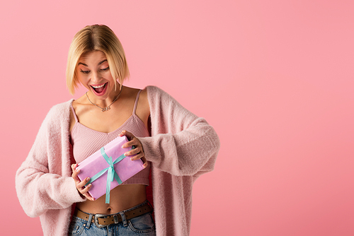 amazed young woman in cardigan holding wrapped gift box isolated on pink