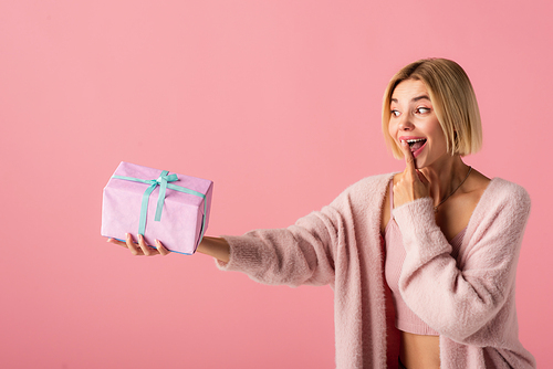 excited young woman in cardigan covering mouth with finger while holding wrapped gift box isolated on pink