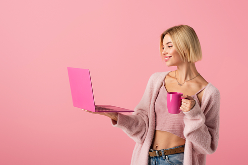 happy woman holding mug with coffee and laptop isolated on pink