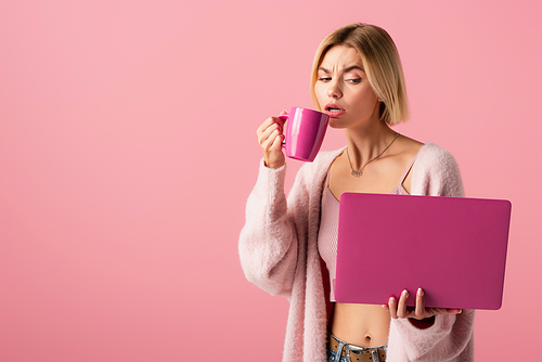 blonde woman holding mug with coffee and laptop isolated on pink