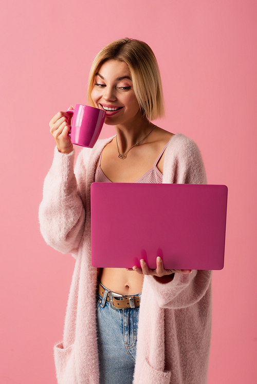 smiling young woman holding mug with coffee and laptop isolated on pink