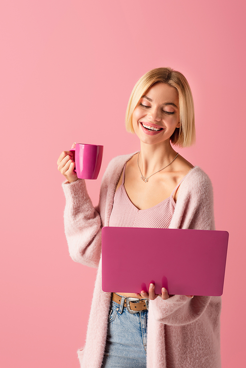 joyful woman holding mug with coffee and laptop isolated on pink