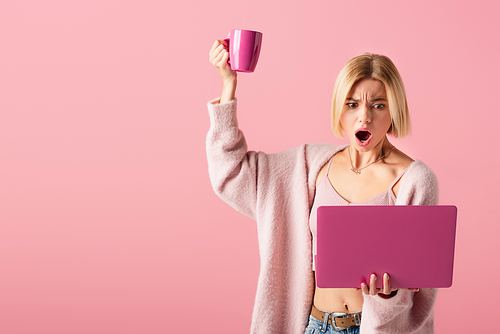 shocked woman holding mug with coffee and laptop isolated on pink