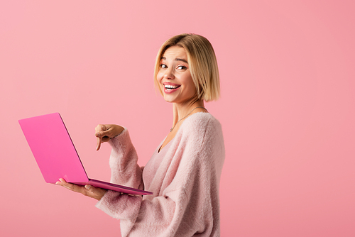 happy young woman pointing with finger at laptop isolated on pink