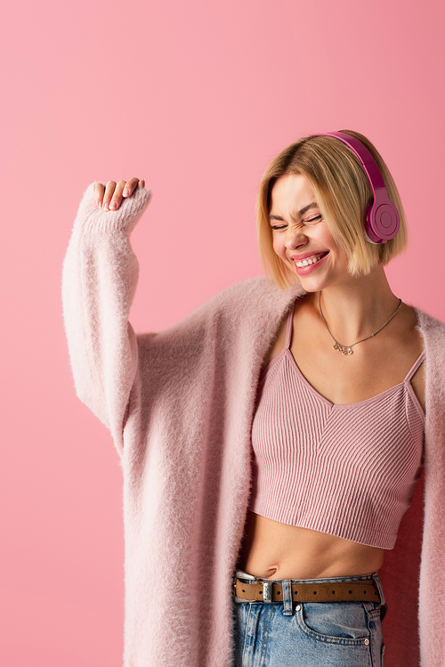 happy blonde woman listening music in wireless headphones isolated on pink