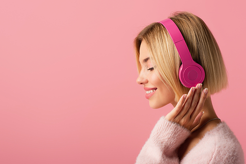 side view of cheerful blonde woman listening music in wireless headphones isolated on pink