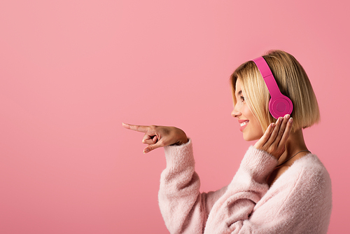 side view of cheerful blonde woman listening music in wireless headphones and pointing with finger isolated on pink