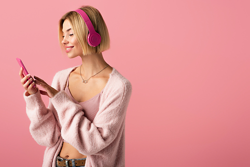 cheerful woman listening music in wireless headphones and using smartphone isolated on pink