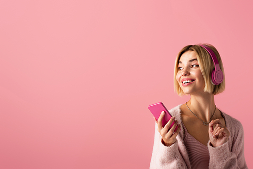 cheerful woman listening music in wireless headphones and holding smartphone isolated on pink