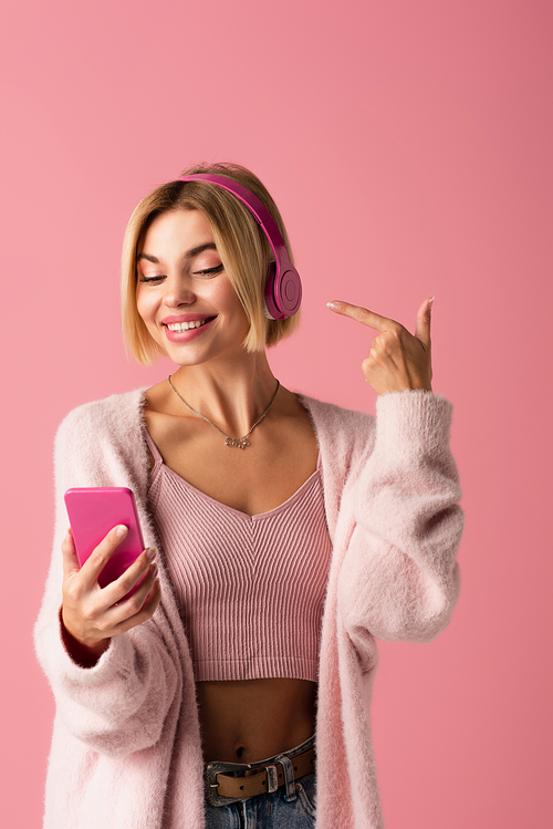 cheerful woman pointing at wireless headphones and holding smartphone isolated on pink