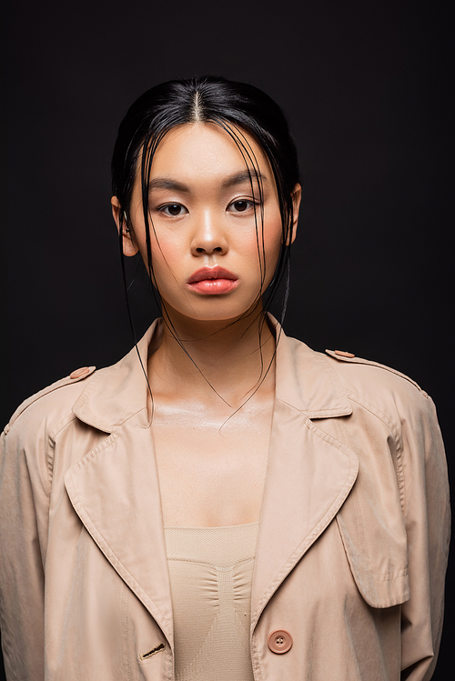 Portrait of asian woman in trench coat looking at camera isolated on black