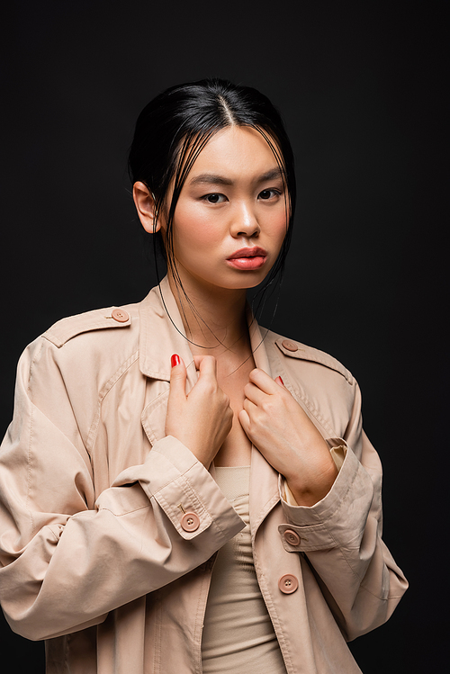 Fashionable asian woman touching trench coat isolated on black