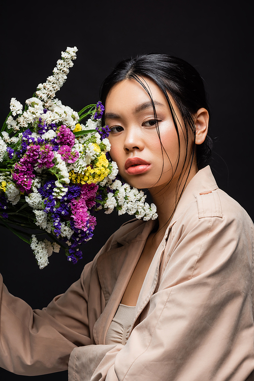 Young asian model in trench coat holding wildflowers near face isolated on black