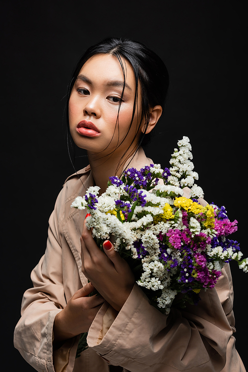 Stylish asian woman in trench coat holding wildflowers isolated on black