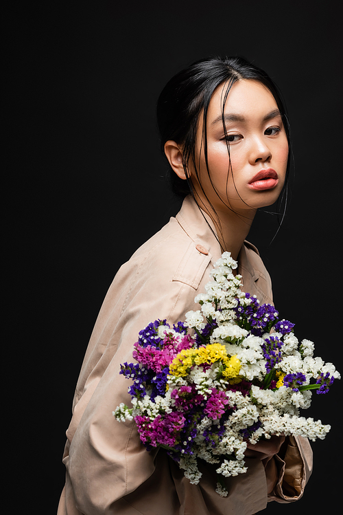 Asian woman in beige trench coat holding colorful wildflowers isolated on black