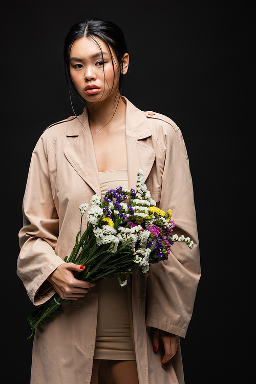 Stylish asian woman in trench coat and beige dress holding flowers isolated on black