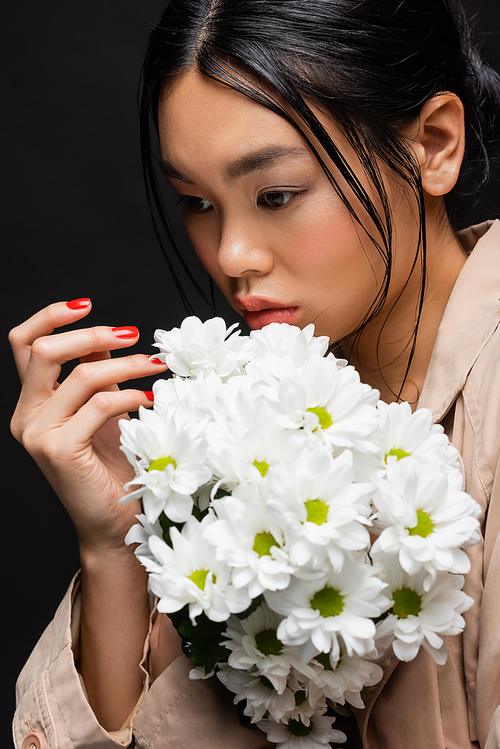 Stylish asian woman in trench coat posing with bouquet of chrysanthemums isolated on black