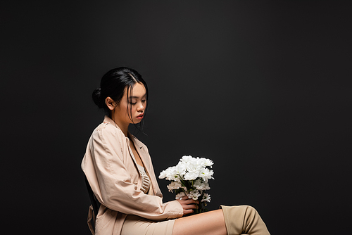 Fashionable asian model in beige dress and trench coat holding white chrysanthemums isolated on black