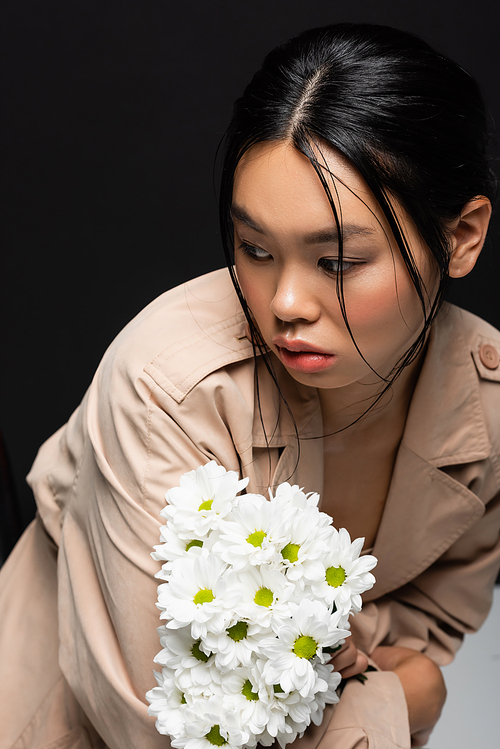 Trendy asian woman in trench coat holding white flowers and looking away on black background