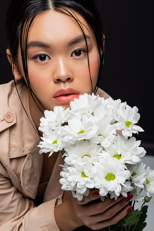 Portrait of asian woman in beige trench coat holding flowers and looking at camera isolated on black