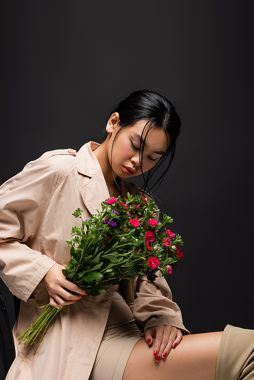 Young asian model in dress and trench coat looking at bouquet isolated on black